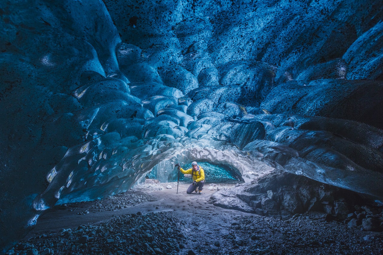 Une personne en veste jaune explore la grotte de glace de Katla, entourée de formations de glace bleutée et de sol rocheux.
