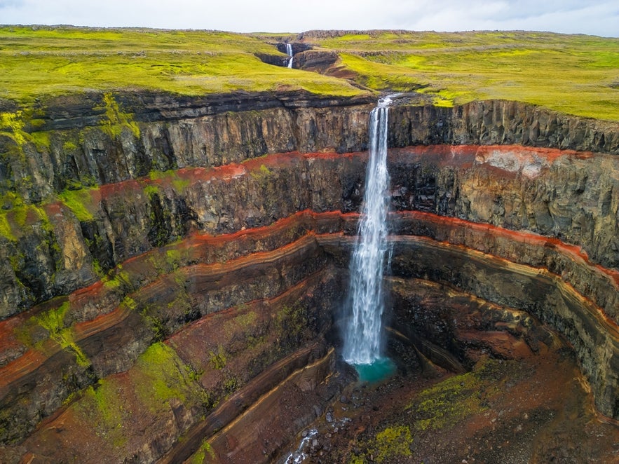 Hengifoss Waterfall plunges down colorful, layered cliffs with red strata and green moss in East Iceland.