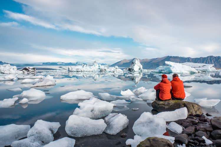 赤いジャケットを着た2人が、ヨークルスアゥルロゥン氷河湖の氷河と静寂に包まれる景色を堪能