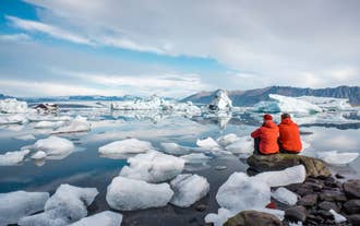 To personer i røde jakker sitter på en stein ved Jökulsárlón isbrelagune, omgitt av flytende isfjell og stille vann.