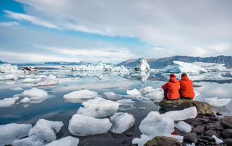 两名身穿红色夹克的游客坐在杰古沙龙冰湖（Jokulsarlon Glacier Lagoon）石上，周围环绕冰山与平静湖面。