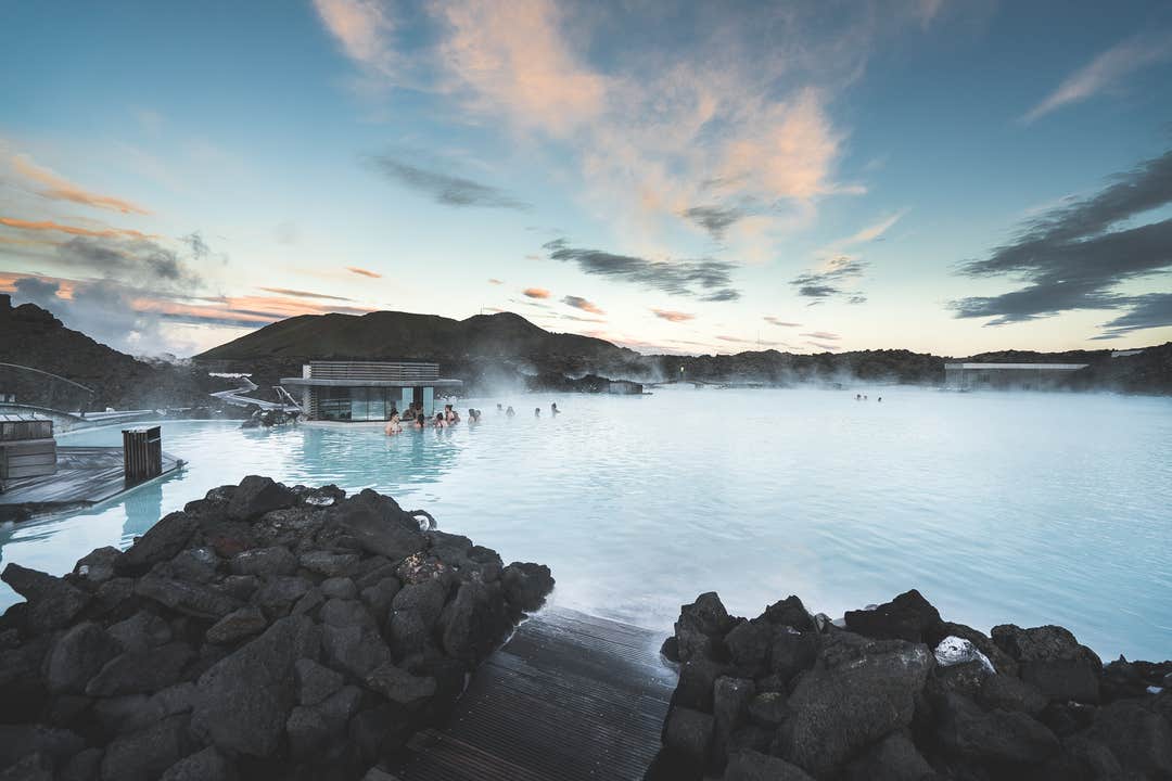 People relax in the steaming Blue Lagoon with black lava rocks and a modern spa building in the background at dusk.