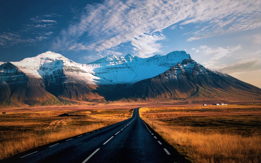 Scenic view of Iceland’s Ring Road leading toward snow-capped mountains under a dramatic sky, surrounded by golden fields.