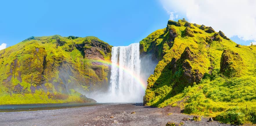 Cascada Skogafoss con un arcoíris en Islandia en julio, rodeada de acantilados verdes bajo un cielo azul brillante. Cascada Skogafoss con un arcoíris en Islandia en julio, rodeada de acantilados verdes bajo un cielo azul brillante.