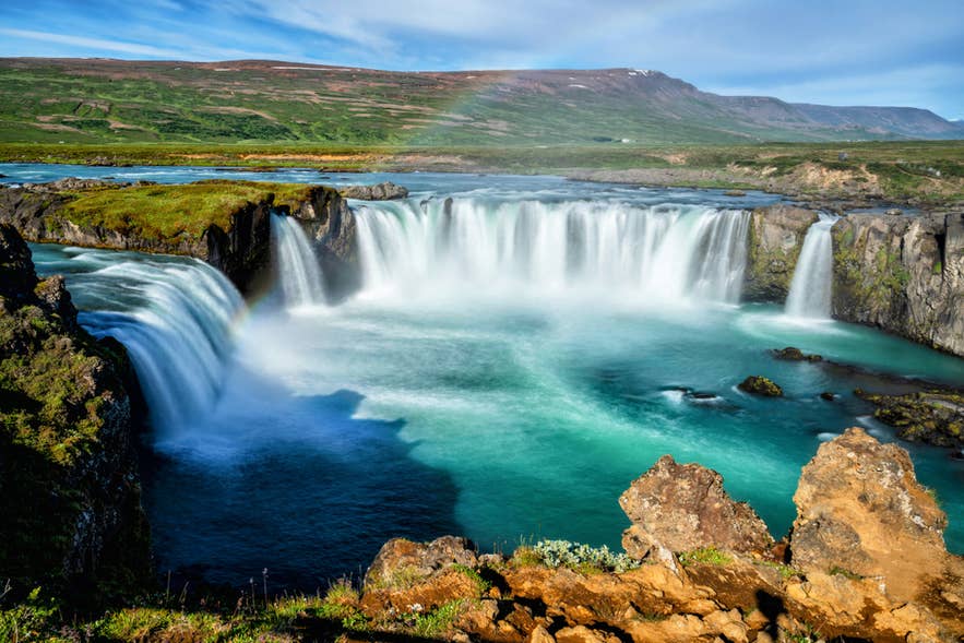 Cascada Godafoss en el norte de Islandia en julio con agua turquesa, colinas verdes y un arcoíris tenue bajo cielos azules.