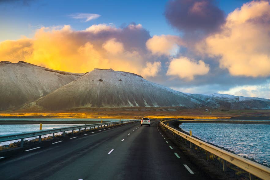 Auto che percorre la Ring Road islandese con montagne, lago e cielo all’alba sullo sfondo.