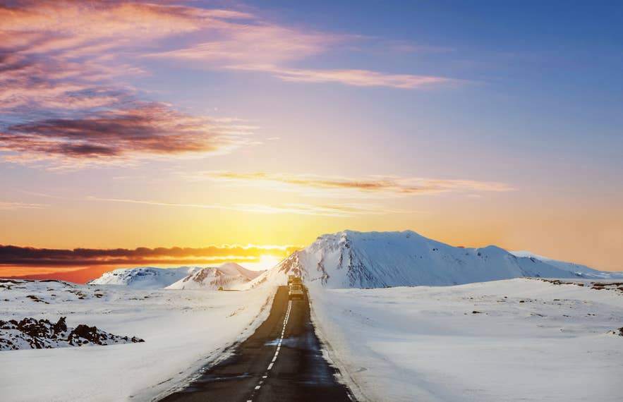 Strada innevata tra le montagne islandesi al tramonto, con un veicolo che si dirige verso l’orizzonte luminoso.
