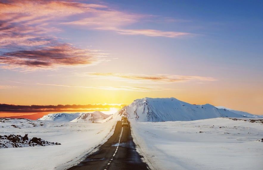 Snow-covered road through Icelandic mountains at sunset, with a vehicle driving toward the glowing horizon.