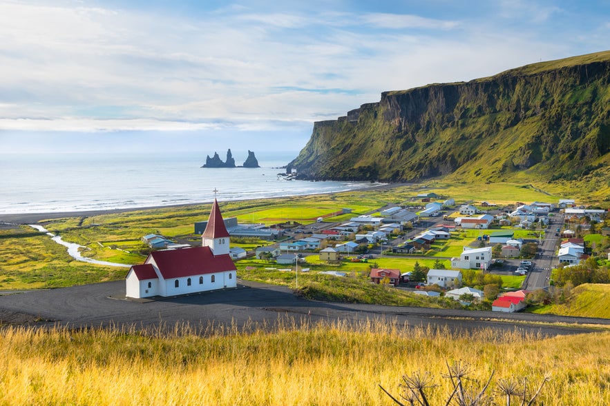 El pueblo de V&iacute;k con su iglesia de techo rojo, playa de arena negra, formaciones rocosas y acantilados bajo un cielo azul.