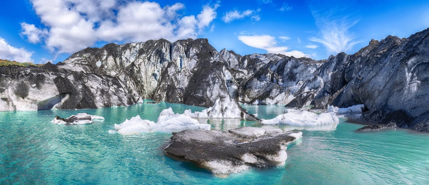 Solheimajokull Glacier with turquoise meltwater and ice chunks under a partly cloudy blue sky.