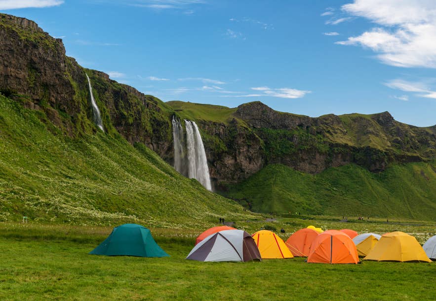 Camping tents near Seljalandsfoss waterfall in southern Iceland, set against lush green cliffs and a bright blue sky.