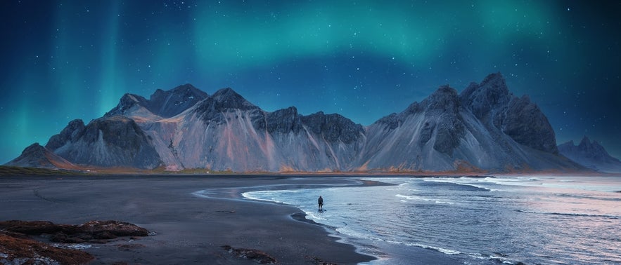 Stokksnes Beach at night with a lone figure, rugged mountains, and northern lights in the sky.