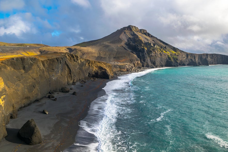 Volcanic black sand beach with cliffs and Festarfjall mountain under cloudy sky by turquoise sea.