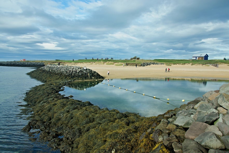 Nautholsvik Beach in Reykjavik with rocky breakwaters, calm water pool, sandy shore, and cloudy sky above.