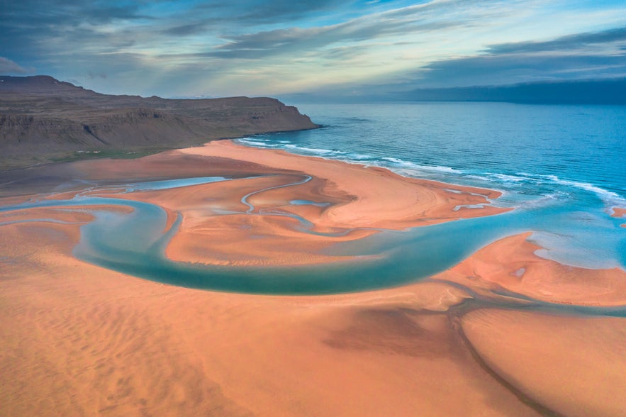 Raudasandur Beach in the Westfjords with orange sand, winding blue water, cliffs, and a cloudy sky.