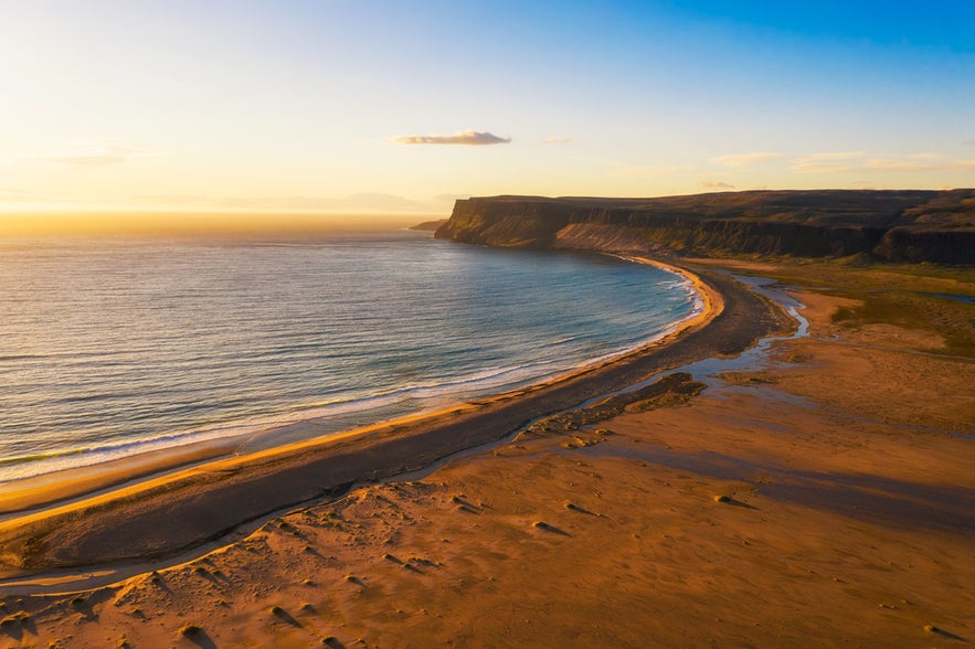 Breidavik Beach at sunset with golden sand, calm waves, and cliffs in the background under a clear sky.