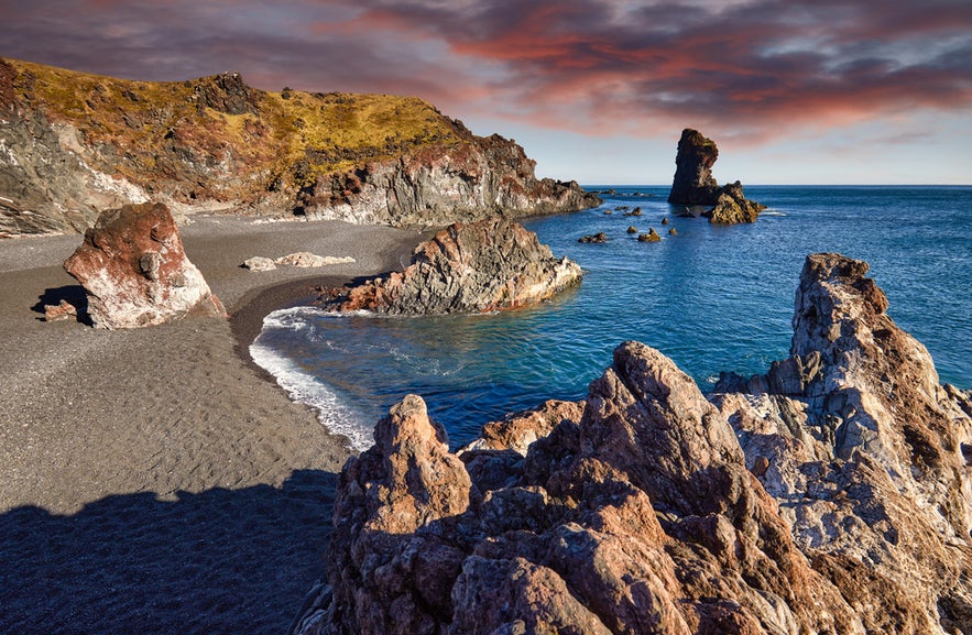 Rocky shoreline and black sand at Djupalonssandur Beach, with sea stacks and a dramatic sunset sky over the ocean.