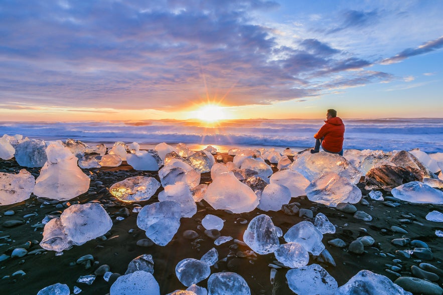 Person sitting among glistening ice chunks on Jokulsarlon Diamond Beach, Iceland, watching the sunrise over the ocean.