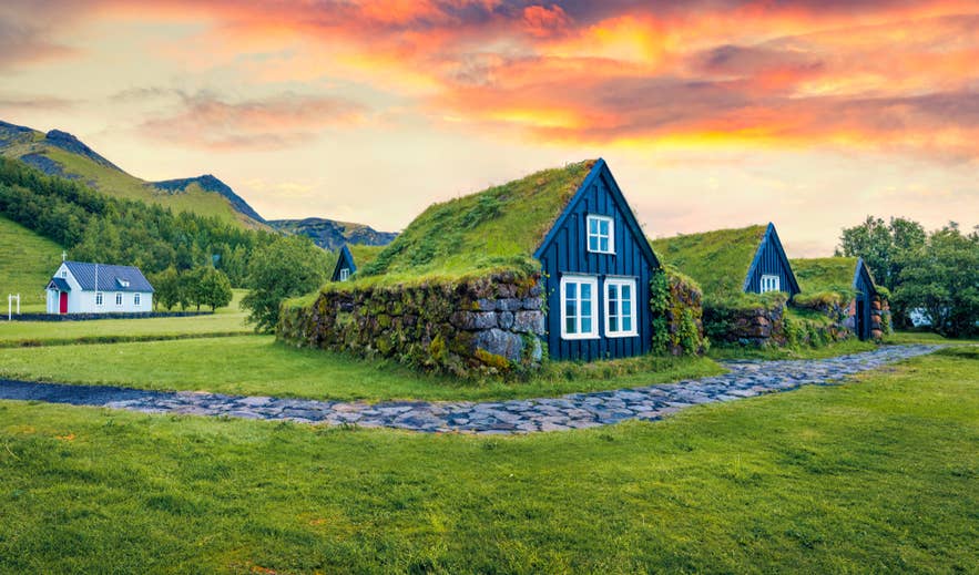 Green turf-roofed houses at Skogar under a colorful summer sunset, with a small white church behind.