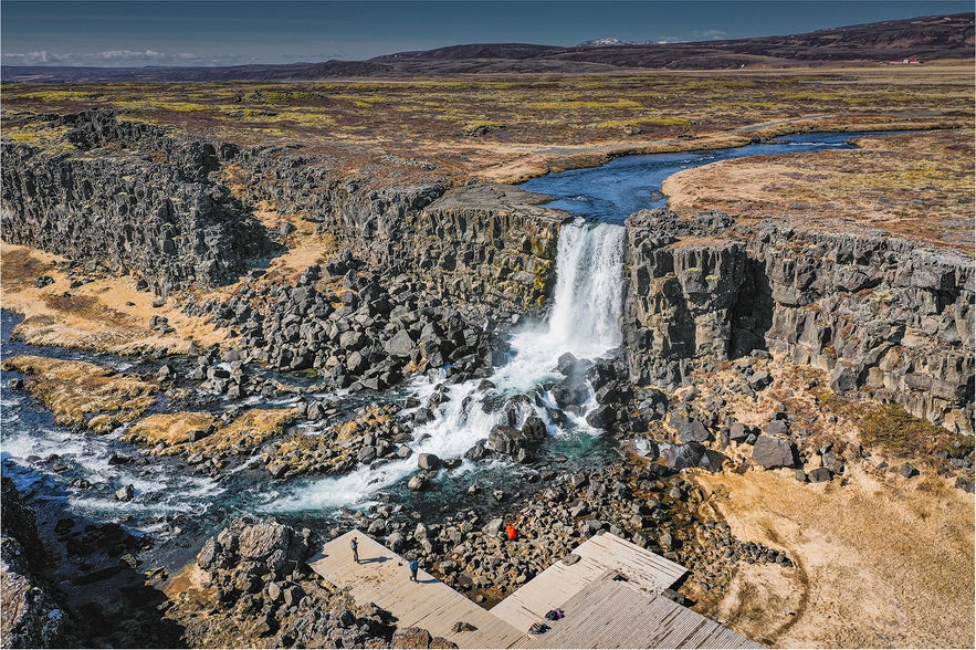 Aerial view of Oxararfoss waterfall flowing over rocky cliffs into a stream, with wooden viewing platforms and visitors nearby.