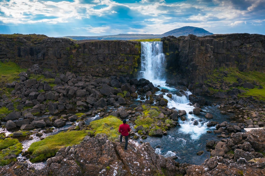 Hiker in red jacket overlooking Oxararfoss waterfall, surrounded by lava rocks, moss, and a flowing stream in Thingvellir.