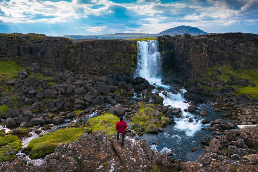 Escursionista con giacca rossa che osserva la cascata Oxararfoss, circondata da rocce laviche, muschio e un torrente nel Parco Nazionale di Thingvellir.
