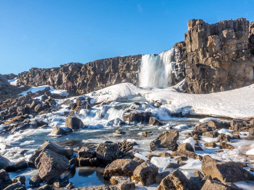 La cascata Oxararfoss in inverno, circondata da rocce e ghiaccio coperti di neve sotto un cielo azzurro nel Parco Nazionale di Thingvellir.