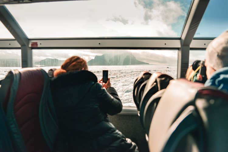 A woman seated at a vehicle window photographing snowy mountains and open Icelandic landscape.