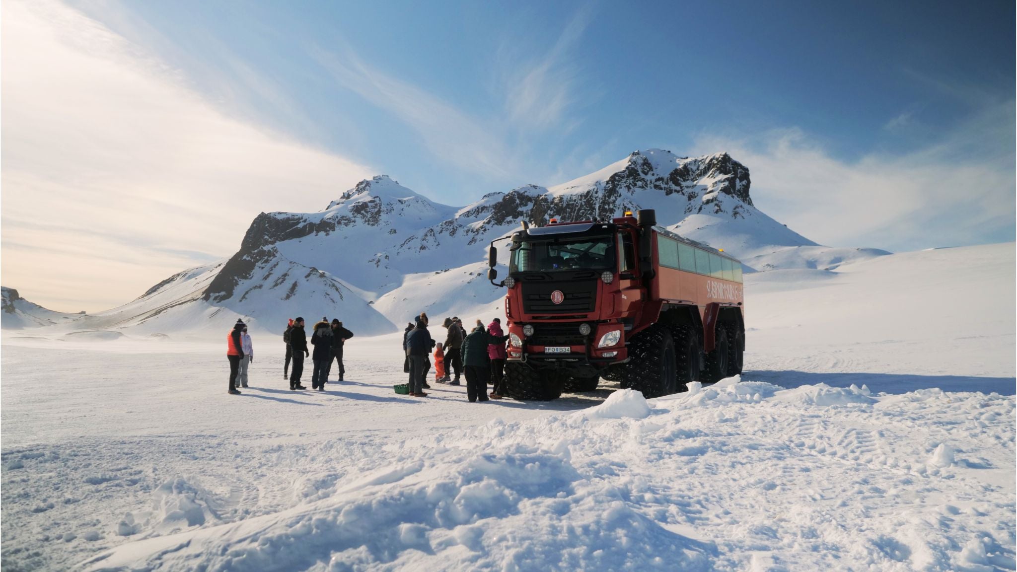 A modified monster truck parked at Langjokull Glacier, surrounded by people and snow-covered mountains.