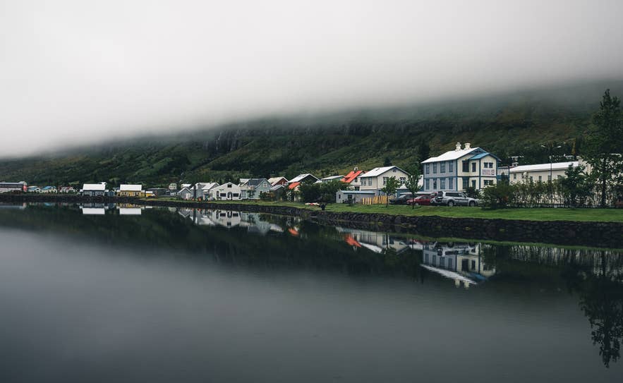 Hus langs en stille fjord under lavt hengende tåke på Island.