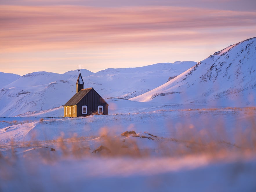 A black wooden church stands in snowy Icelandic hills, lit by soft sunrise light under streaks of pink and orange clouds.