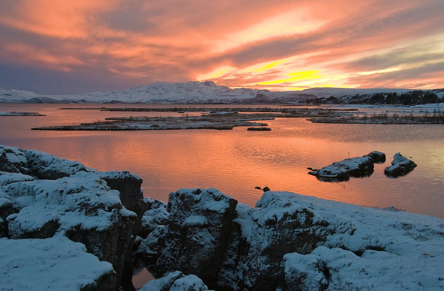 Snow-covered rocks surround a calm lake glowing orange under a dramatic winter sunset in Thingvellir National Park, Iceland.