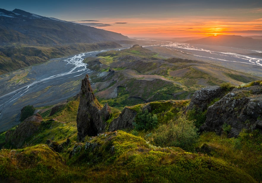 A winding glacial river cuts through mossy mountains and black volcanic sand as the sun sets behind distant ridges in southern Iceland.