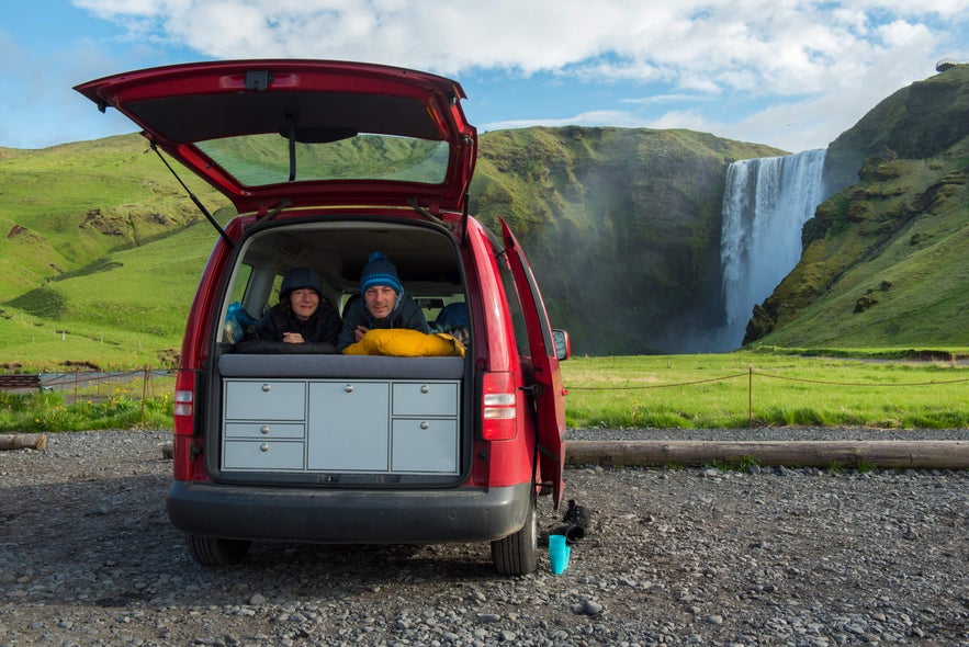 A couple relaxing in a campervan during a scenic camping trip, with Skogafoss Waterfall in the background.