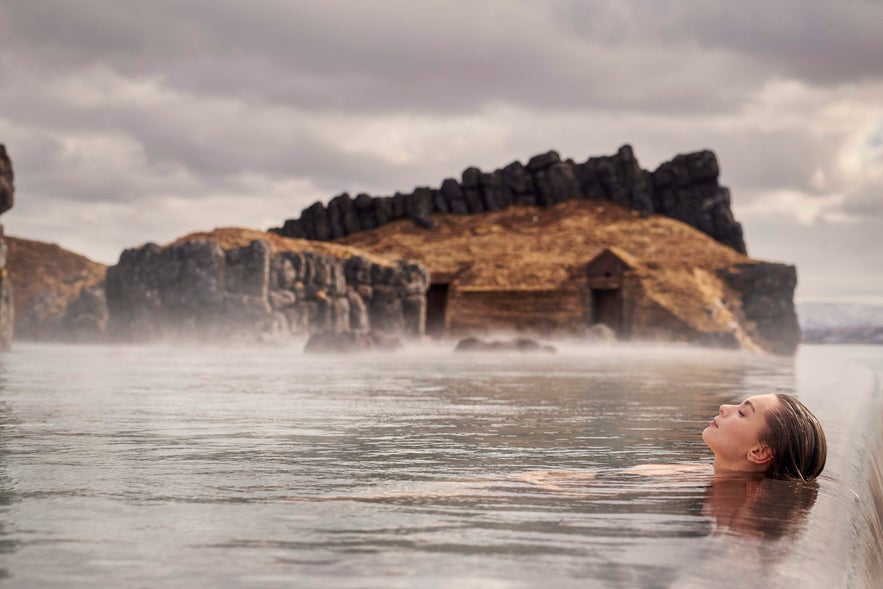 Woman relaxing in the warm waters of Sky Lagoon, Iceland, with dramatic rocky cliffs and turf houses in the background.