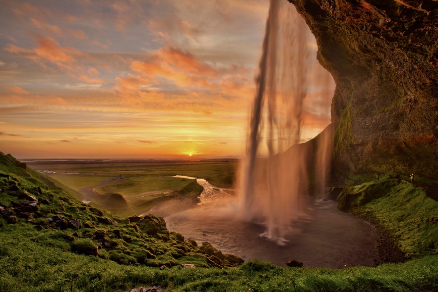 Seljalandsfoss waterfall glowing under the midnight sun in Iceland, with golden skies and lush green hills.