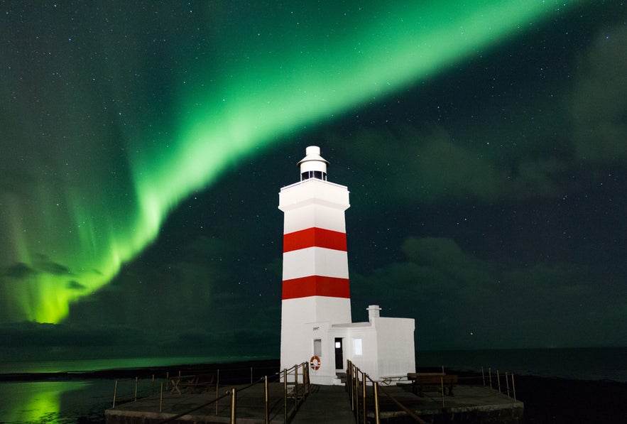 Grotta Lighthouse in Iceland beneath a vibrant green aurora borealis lighting up the night sky over the ocean.