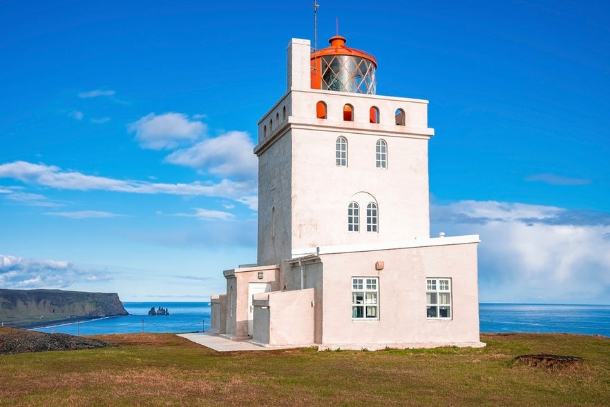 Dyrholaeyjarviti lighthouse in Iceland stands on a cliffside with ocean views and sea stacks in the distance under blue skies.