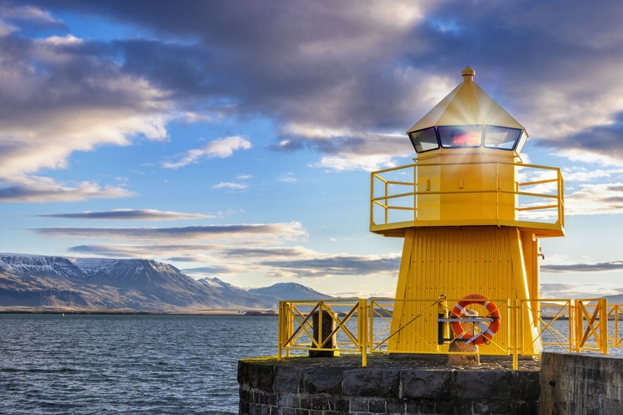 Bright yellow lighthouse at Reykjavik Harbour glowing at sunrise, with snowy mountains and calm waters in the background.