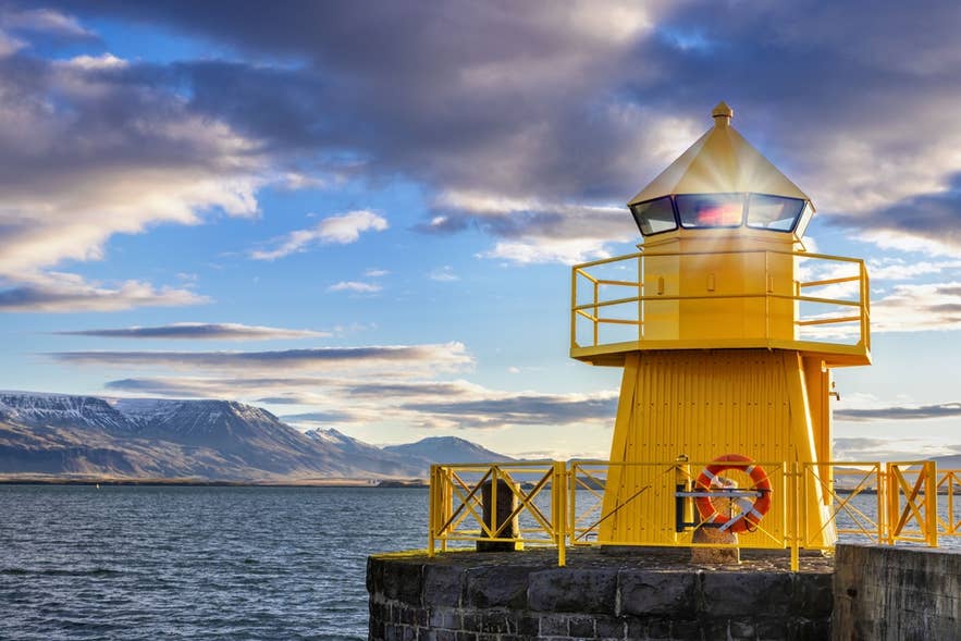 Bright yellow lighthouse at Reykjavik Harbour glowing at sunrise, with snowy mountains and calm waters in the background.