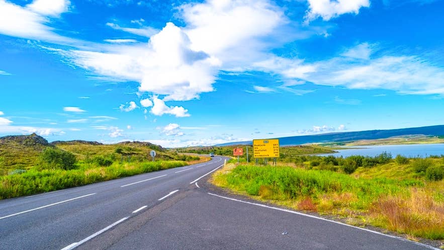 A rural paved road passes yellow distance signs by a lake in North Iceland on a calm summer day.