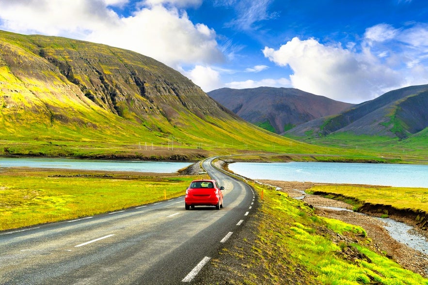 Red car driving in Iceland in August on a scenic fjord road with mountains and blue skies in the Westfjords Region.