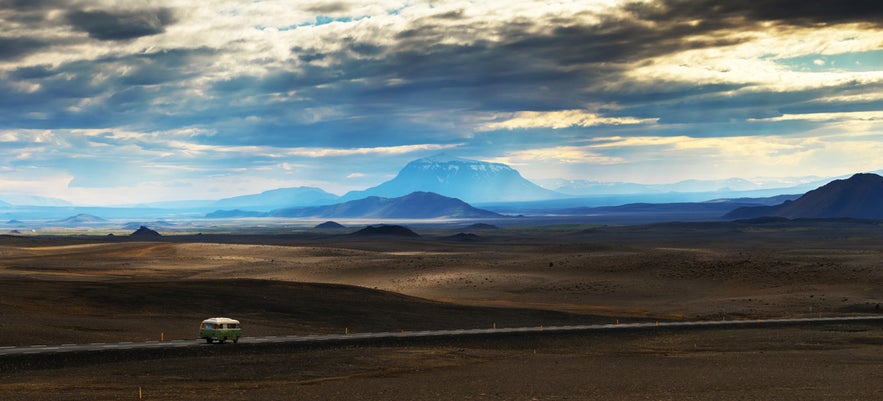 A van drives through Iceland's highlands with Herdubreid mountain rising in the distance under a dramatic, cloudy sky.