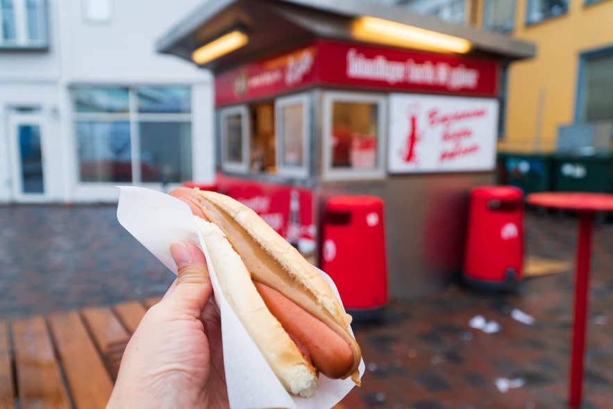 Hand holding an Icelandic hot dog with sauce in front of the famous Baejarins Beztu Pylsur red hot dog stand in Reykjavik. Hand holding an Icelandic hot dog with sauce in front of the famous Baejarins Beztu Pylsur red hot dog stand in Reykjavik.