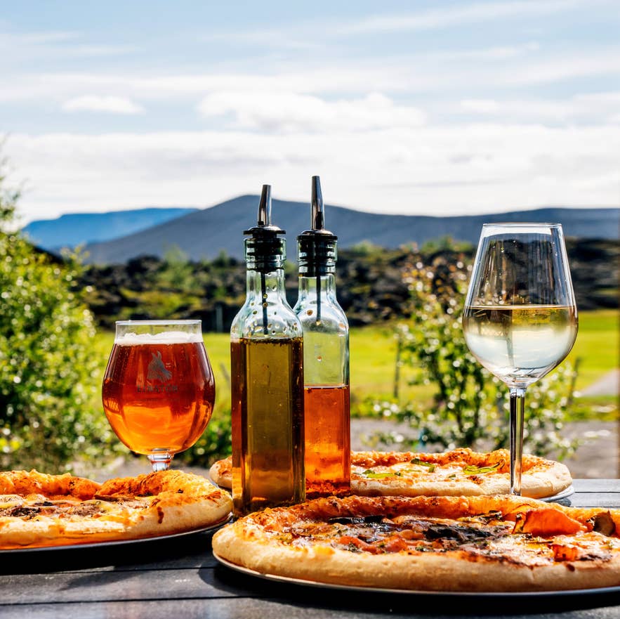 Pizzas with beer, white wine, and oil bottles on an outdoor table overlooking green fields and volcanic hills under a blue sky.