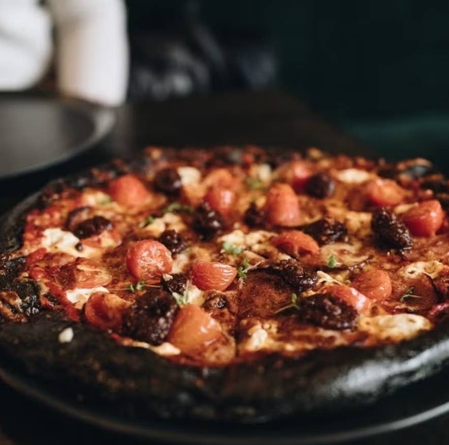 Pizza à croûte couleur charbon garnie de tomates cerises, de saucisse et de fromage, servie sur une assiette noire dans une ambiance de restaurant tamisée.
