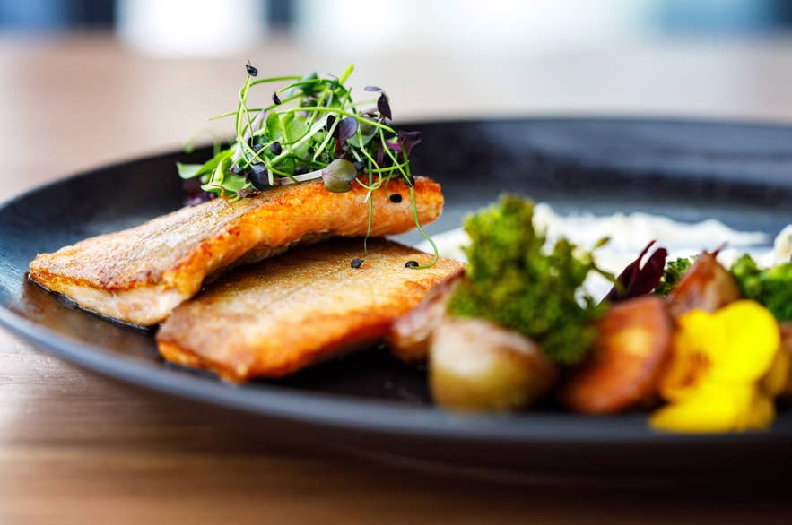 Filets de poisson poêlés garnis de pousses, servis avec des pommes de terre rôties, du brocoli et des fleurs comestibles sur une assiette noire.
