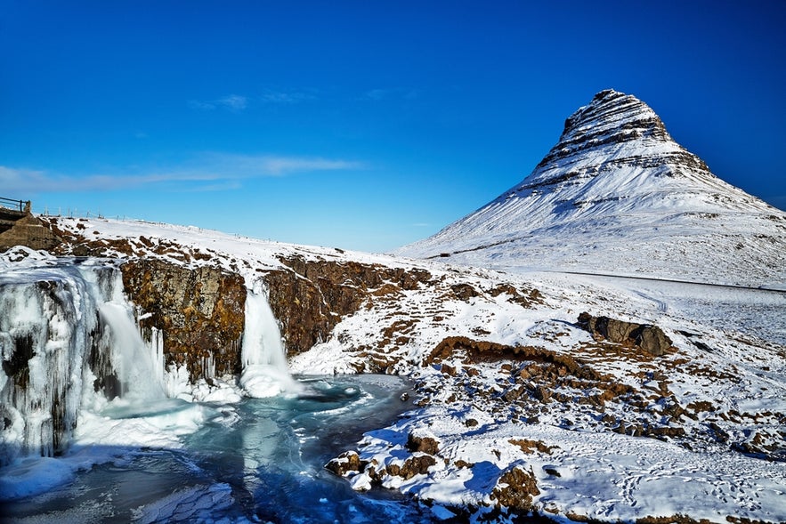 Snow-covered Kirkjufell mountain and frozen Kirkjufellsfoss waterfall under a clear blue sky in Iceland's winter landscape.