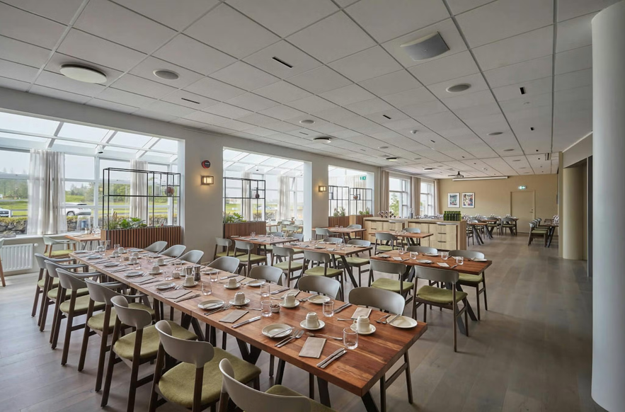 Bright, modern dining room with wooden tables, neatly set for service, surrounded by large windows and natural light.