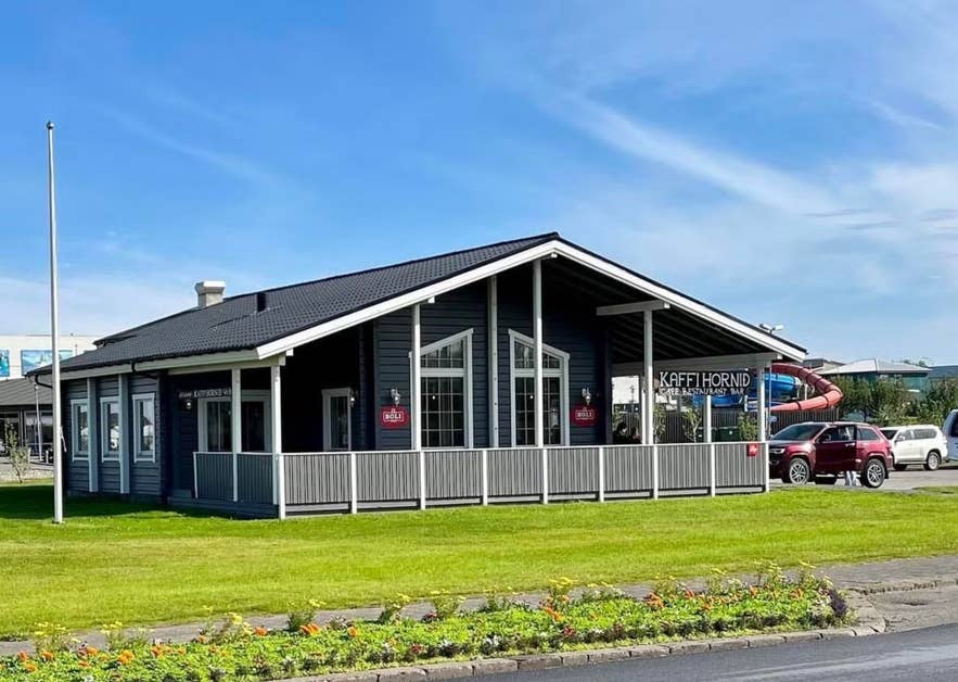 Dunkelgraues Holzgebäude, in dem sich das Kaffi Hornið Café, Restaurant und Bar befindet, mit einer Veranda vorne und geparkten Autos bei blauem Himmel.